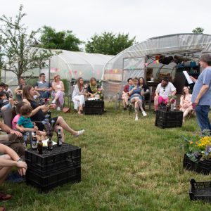 People gathered outside a greenhouse, engaged in conversation and handling plants