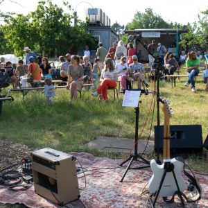 A lively outdoor gathering with people seated on the grass, a stage with speakers, and musical equipment