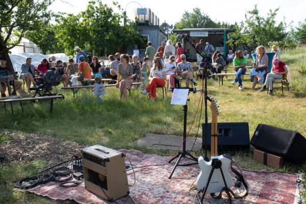 A lively outdoor gathering with people seated on the grass, a stage with speakers, and musical equipment