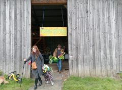 Two people carrying baskets of fresh produce while exiting a wooden barn with a yellow sign.